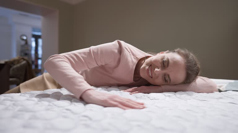 Close-up white orthopedic mattress with smiling elegant woman lying down caressing soft comfortable bedding. Portrait of happy Caucasian female buyer choosing cozy bed in furniture store showroom.