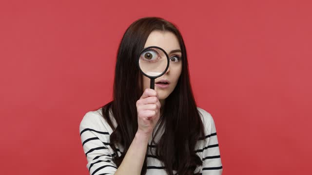 Portrait of woman looks into camera through magnifying glass, inspects something, private detective.