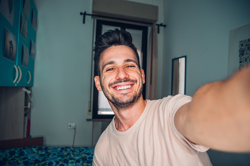 Happy young man taking a selfie portrait with smart phone mobile at home - Smiling guy looking at camera - Video blogger recording new video streaming indoor