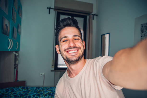 jovem feliz tirando um retrato de selfie com celular de smartphone em casa - cara sorridente olhando para a câmera - blogueiro de vídeo gravando novo vídeo streaming dentro de casa - mobile phone home video camera video photograph - fotografias e filmes do acervo