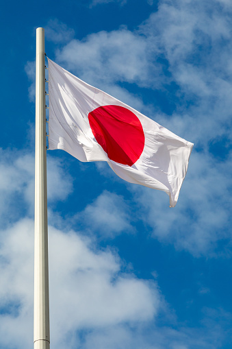 The flag of Japan flutters on a flagpole against the sky with clouds. Vertical