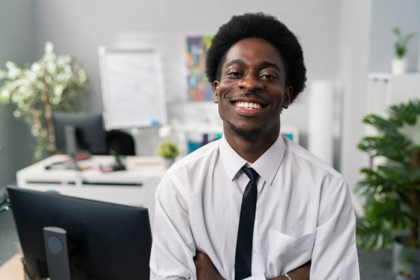 Smiling friendly dark skinned afro man sits on desk wearing white shirt and tie, in background scenery of company, office boss, president, manager, businessman, arms crossed on stomach