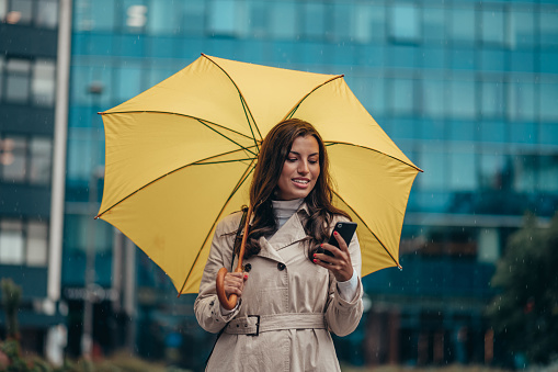 Beautiful young woman using smartphone and holding a yellow umbrella during a heavy rain in the city