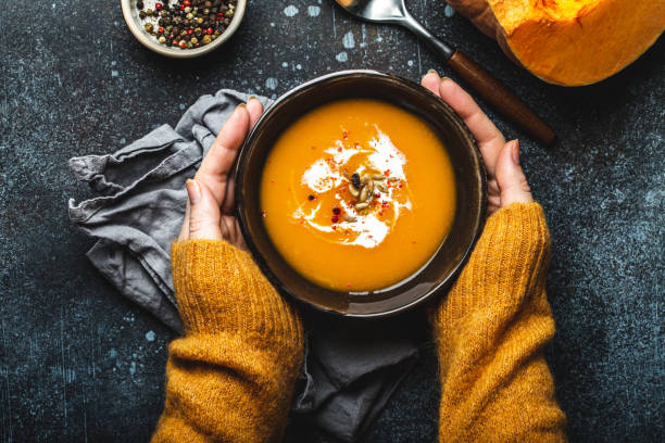 Female hands with bowl of pumpkin soup Female hands in yellow knitted sweater holding a bowl with pumpkin cream soup on dark stone background with spoon decorated with cut fresh pumpkin, top view. Autumn cozy dinner concept soup stock pictures, royalty-free photos & images