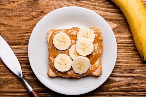 Toast with peanut butter and banana on white ceramic plate with knife on rustic wooden background, top view flat lay