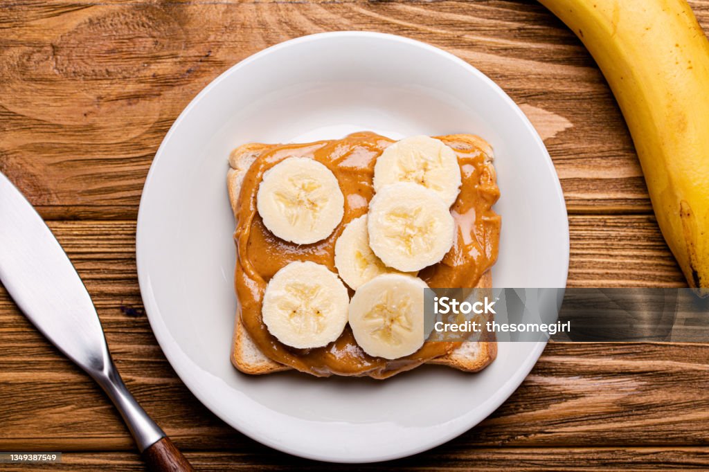 Toast with peanut butter and banana Toast with peanut butter and banana on white ceramic plate with knife on rustic wooden background, top view flat lay Banana Stock Photo