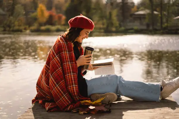 A Teengirl in a red beret reading book on wooden pontoon. Autumn season. Female with trendy clothes relaxing near water reading book and drink coffee.  Autumn mood. Autumn concept. Autumn day.