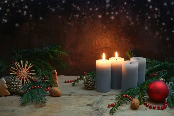 Second Advent, two of four candles are lighted, red bauble, straw star, fir branches and Christmas decoration on rustic wooden planks against a dark brown background, copy space, selected focus
