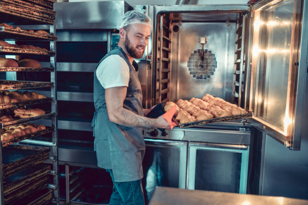 uśmiechnięty brodaty piekarz wyjmuje rogaliki przed spaleniem w piekarniku - chef baker bakery flour zdjęcia i obrazy z banku zdjęć