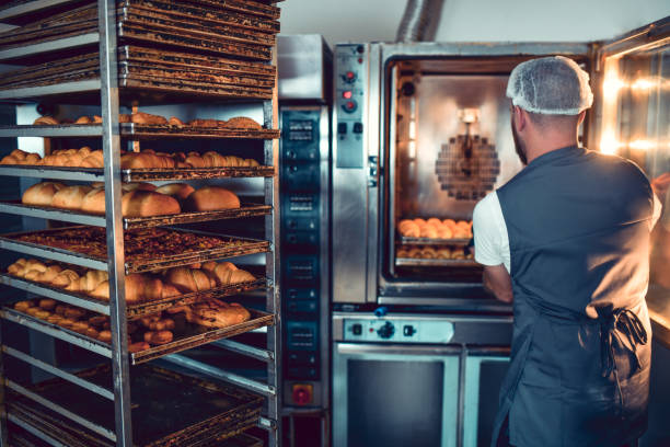 istock Rear View Of Baker With Apron Putting Multiple Trays In Large Baking Oven 1349392203