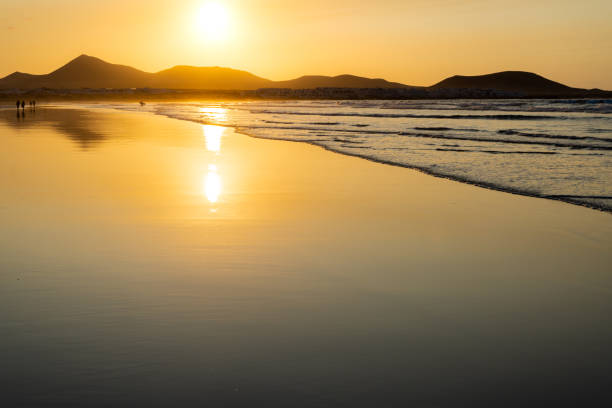 strandlandschaft bei sonnenuntergang mit entfernten surfern in famara strand, lanzarote, kanarische inseln, spanien. - famara stock-fotos und bilder