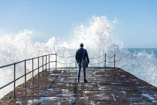 A casual business man carrying a shoulder bag stands on a coastal harbour and looks out to sea as waves crash against the harbour wall.
