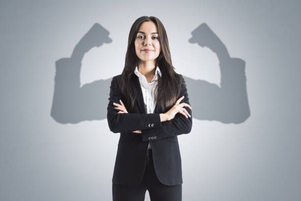 portrait d’une jolie jeune femme d’affaires européenne aux bras croisés et aux bras musclés d’ombre sur fond de mur en béton. force et concept de leadership. - business super hero photos et images de collection