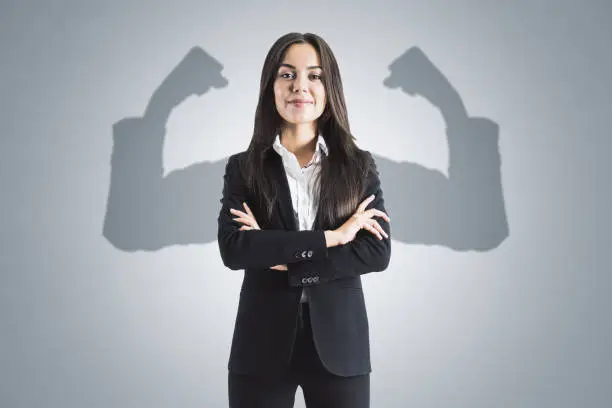 Photo of Portrait of attractive young european businesswoman with folded arms and shadow muscle arms on concrete wall background. Strenght and leadership concept.