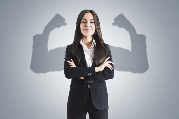 portrait of attractive young european businesswoman with folded arms and shadow muscle arms on concrete wall background. strenght and leadership concept. - superhero imagens e fotografias de stock