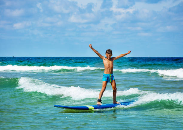 Young 10 years old boy posing with lifted hands ride surfboard on a wave in the sea