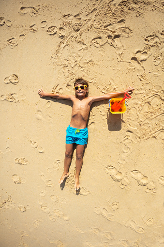 Happy 10 years old boy laying relaxed on the beach with bucket and shovel view from above