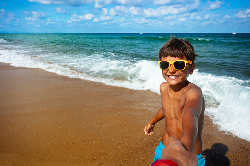 Cute little boy in sunglasses pull parent's hand to the sea with big smile and waves on background