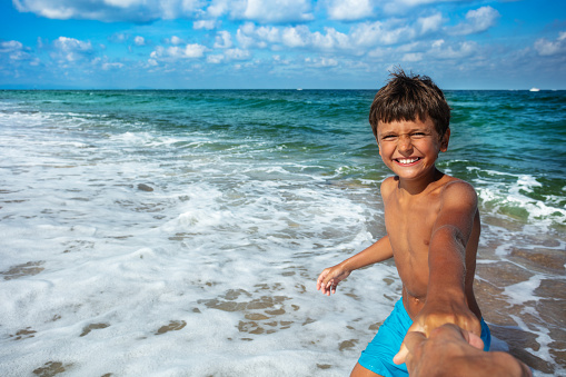 Happy little boy smiling pull parent's hand to the sea with big smile and waves on background