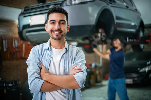 Satisfied male customer at the car garage facing camera with arms crossed while mechanic checks the wheels of the car at background - Automobile industry concepts