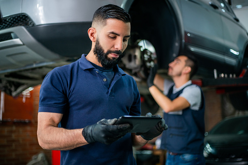 Supervisor at a car workshop checking tablet while mechanic works at background on a car