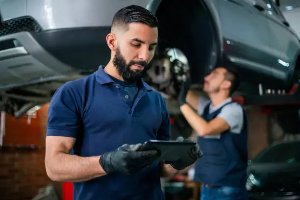 Photo of Supervisor at a car workshop checking tablet while mechanic works at background on a car