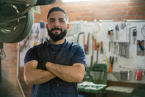 Latin American strong mechanic working at an auto repair shop facing camera with arms crossed while smiling  - car industry concepts