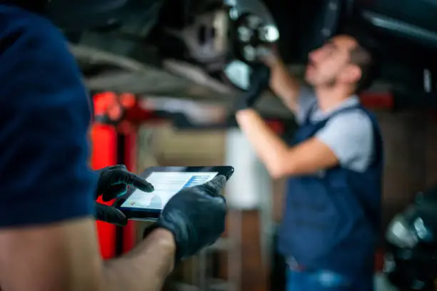 Photo of Focus on foreground of engineer holding a tablet checking the readings of the carâs sensor while the mechanic adjusts them on the disk