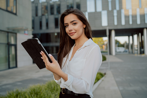 Attractive pretty girl in white shirt stands in front of glass modern business building of insurance corporation company, woman holds tablet in hand browses web writes email reads news