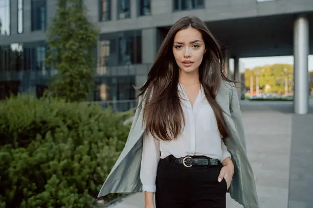 Photo of Attractive girl with brown hair walks out of corporate building for break, dressed in smart shirt and jacket imposed on back, walks outside past glass skyscraper, secretary, businesswoman, boss