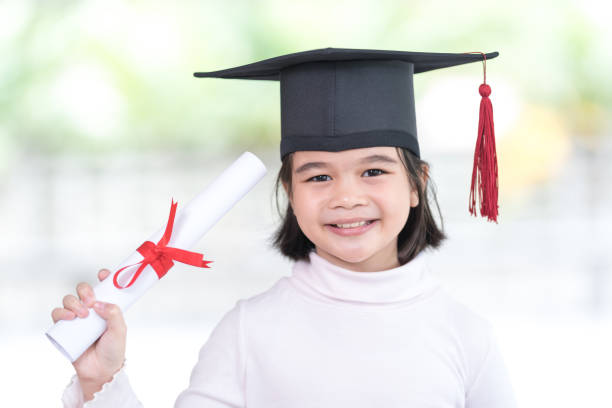 Portrait happy Asian female school kid graduate in a graduation cap holds a rolled certificate paper stock photo