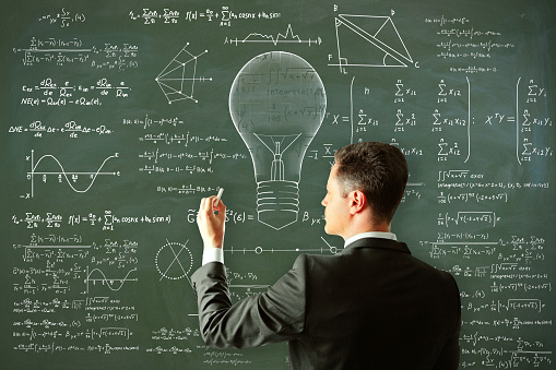 Attractive young european man drawing abstract lamp sketch with mathematical formulas on chalkboard/blackboard wall background. Intelligence, idea, solution, science and innovation concept
