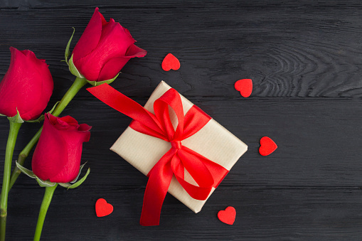 Gift with tied red bow, red roses and red  hearts on the black wooden  background. Top view. Copy space.