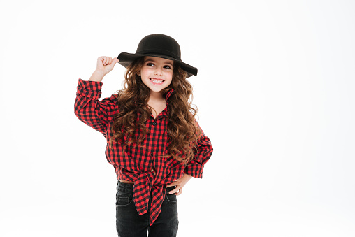 Smiling curly little girl in plaid shirt and hat standing over white background