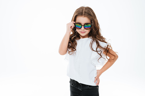 Beautiful curly little girl in sunglasses standing and posing over white background