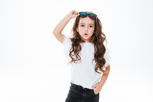 Amazed curly little girl in sunglasses standing and posing over white background