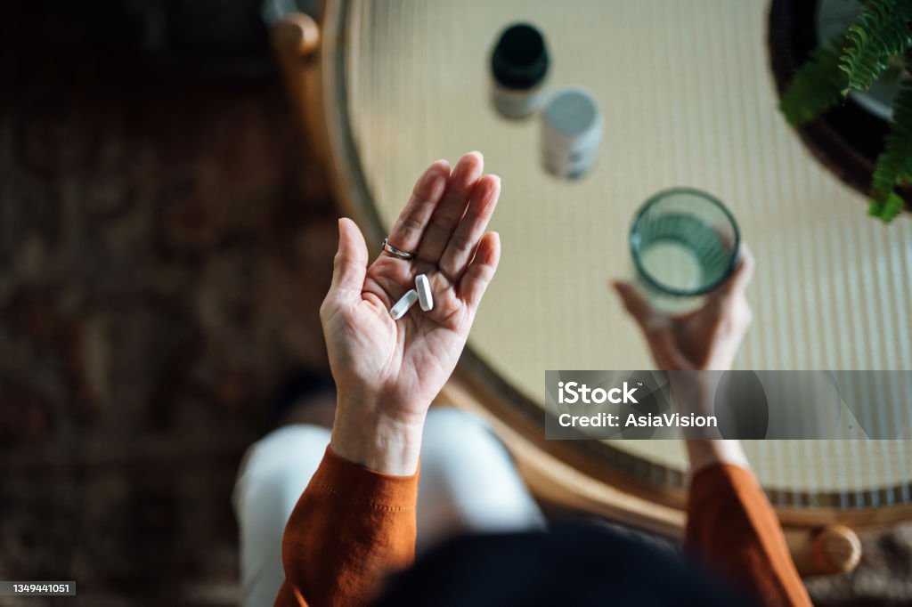 Overhead view of senior Asian woman feeling sick, taking medicines in hand with a glass of water at home. Elderly and healthcare concept Capsule - Medicine Stock Photo
