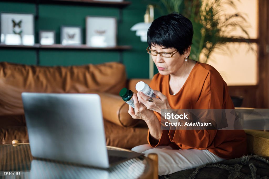 Senior Asian woman having a virtual appointment with doctor online, consulting her prescription and choice of medication on laptop at home. Telemedicine, elderly and healthcare concept Medicine Stock Photo