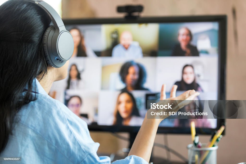 Rear view woman leading virtual meeting with multi-ethnic group The focus of the photo is on the unrecognizable woman in the foreground.  She is gesturing while meeting virtually with an unrecognizable group of diverse co-workers Virtual Event Stock Photo