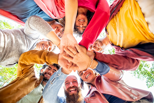directly below portrait of happy diverse large group of multicultural friends holding hands making high five stacking them together outdoors. convivial people having fun. friendship, lifestyle concept