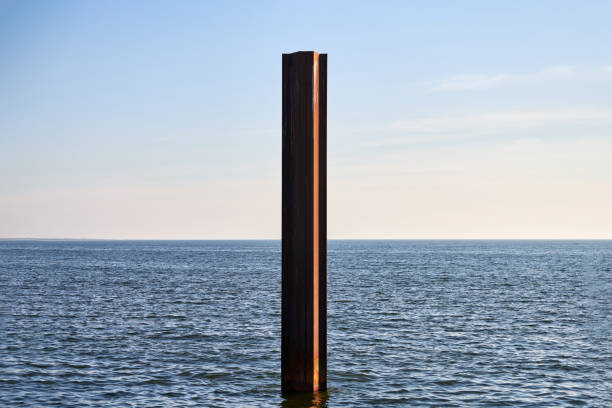 Large rusty steel construction pillar in blue sea with calm waves. Vertical abandoned building part, construction site near coastline. Ecology and recycling concept