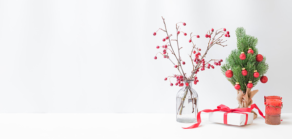 Christmas, New Year home decor. Branches with red berries in a glass vase, christmas tree, gift box on a light background. Mock up for displaying works