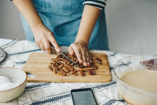 Woman chopping pecan nuts for a cake and using smartphone to read the recipe
