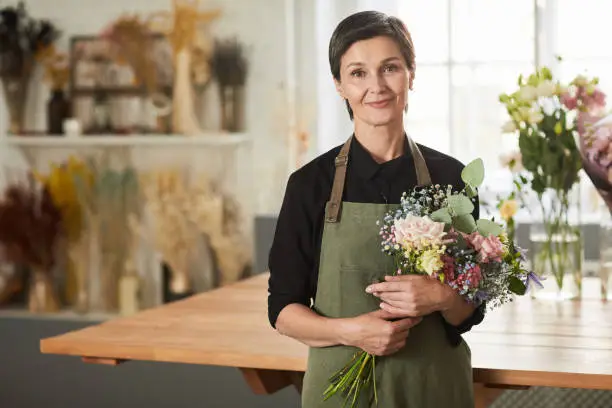 Waist up portrait of smiling adult woman holding flowers in flower shop and looking at camera, copy space
