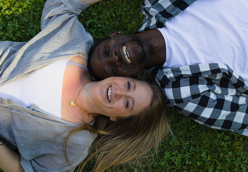 An afro dude and blue-eyed blonde woman lying on the grass while smiling in a public green area.