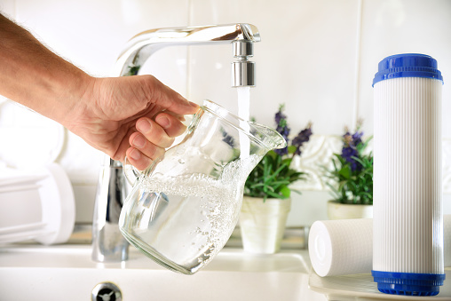 Man filling a glass jug from a tap of purified water with an osmosis system of the home kitchen sink with filters around. Front view. Horizontal composition.
