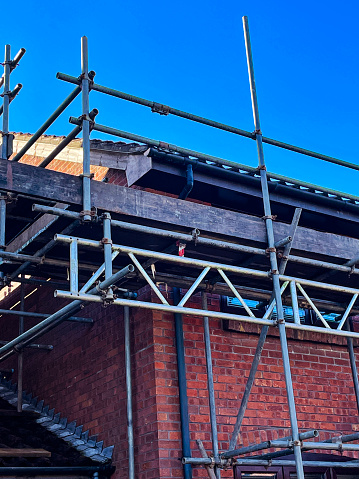 Stock photo showing a close-up view of a modern redbrick house with erected, metal scaffolding poles with the structure held together with clamps or 'couplers', washers, nuts and bolts. The lightweight scaffolding tower features a platform made from wooden boards enabling access for fascia board replacement.