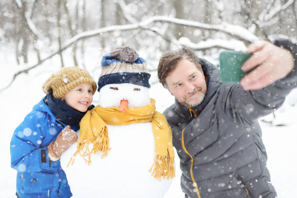 el niño pequeño y su padre tomándose una selfie en el fondo del muñeco de nieve en el parque nevado. ocio activo al aire libre con niños en invierno. niño durante un paseo en un parque de invierno nevado - pareja acogido al aire libre fotografías e imágenes de stock