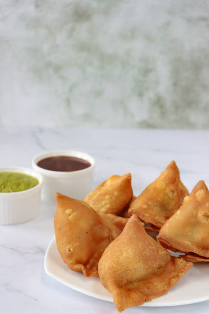 istock Image of five fried samosas on a white plate, stuffed with spiced potato, peas and meat besides ramekins of mint coriander dip and mango chutney, marble effect background, elevated view, focus on foreground, copy space 1349492202