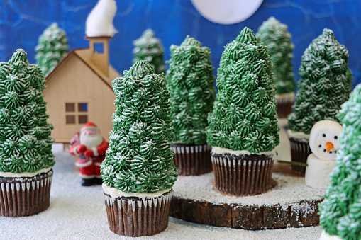 Stock photo showing close-up view of a batch of freshly baked, homemade Christmas tree design chocolate cupcakes in a snowy, night conifer forest scene with a marshmallow snowman and model house with Father Christmas.